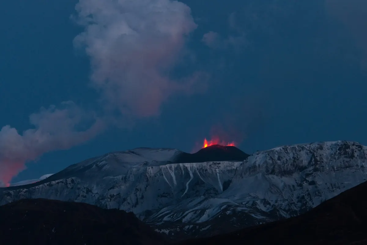 Ein kleiner, im Dämmerlicht rot glühender Krater auf einem schneebedeckten Gebirgspass, daneben eine graue Dampfwolke, die aufsteigt