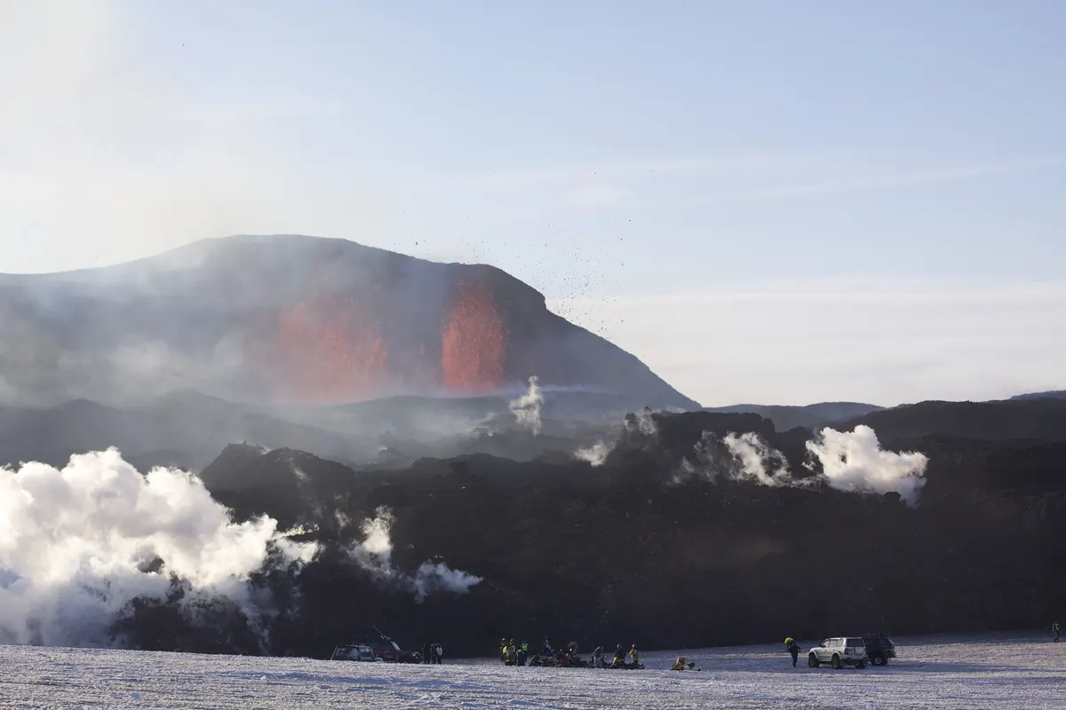 黒い尾根から噴き上がる赤い溶岩の噴水。その下には白い蒸気の雲が広がり、手前の雪の上には数人が立っている