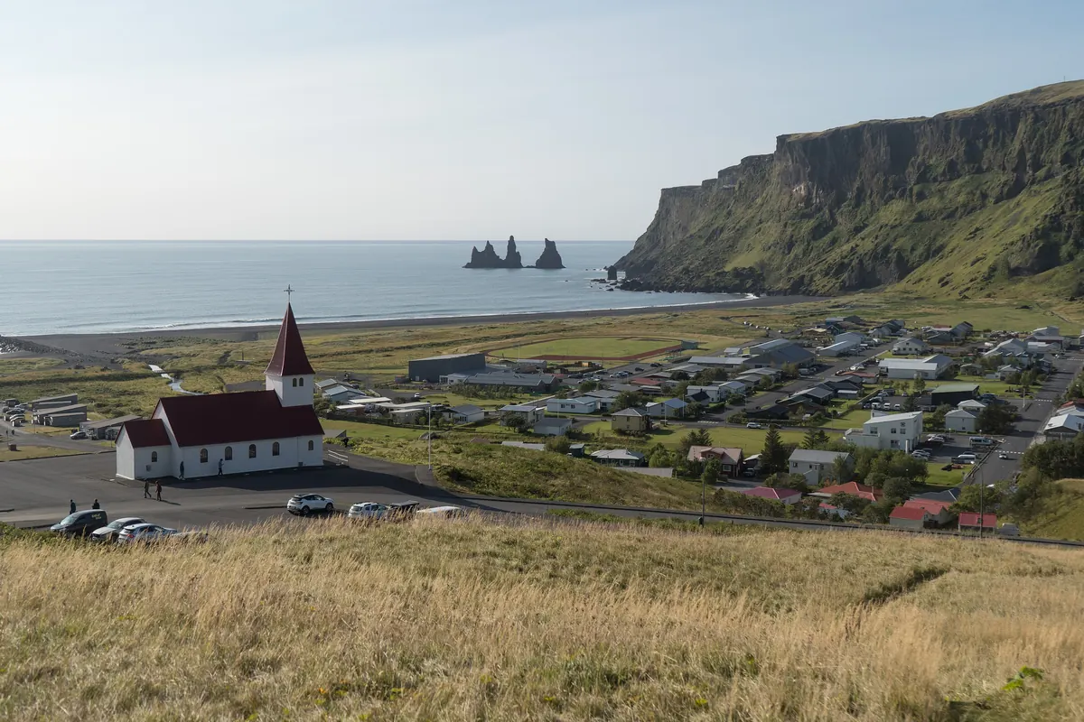La iglesia blanca de tejado rojo sobre el pueblo de Vík, con una playa de arena negra y tres farallones marinos a sus espaldas