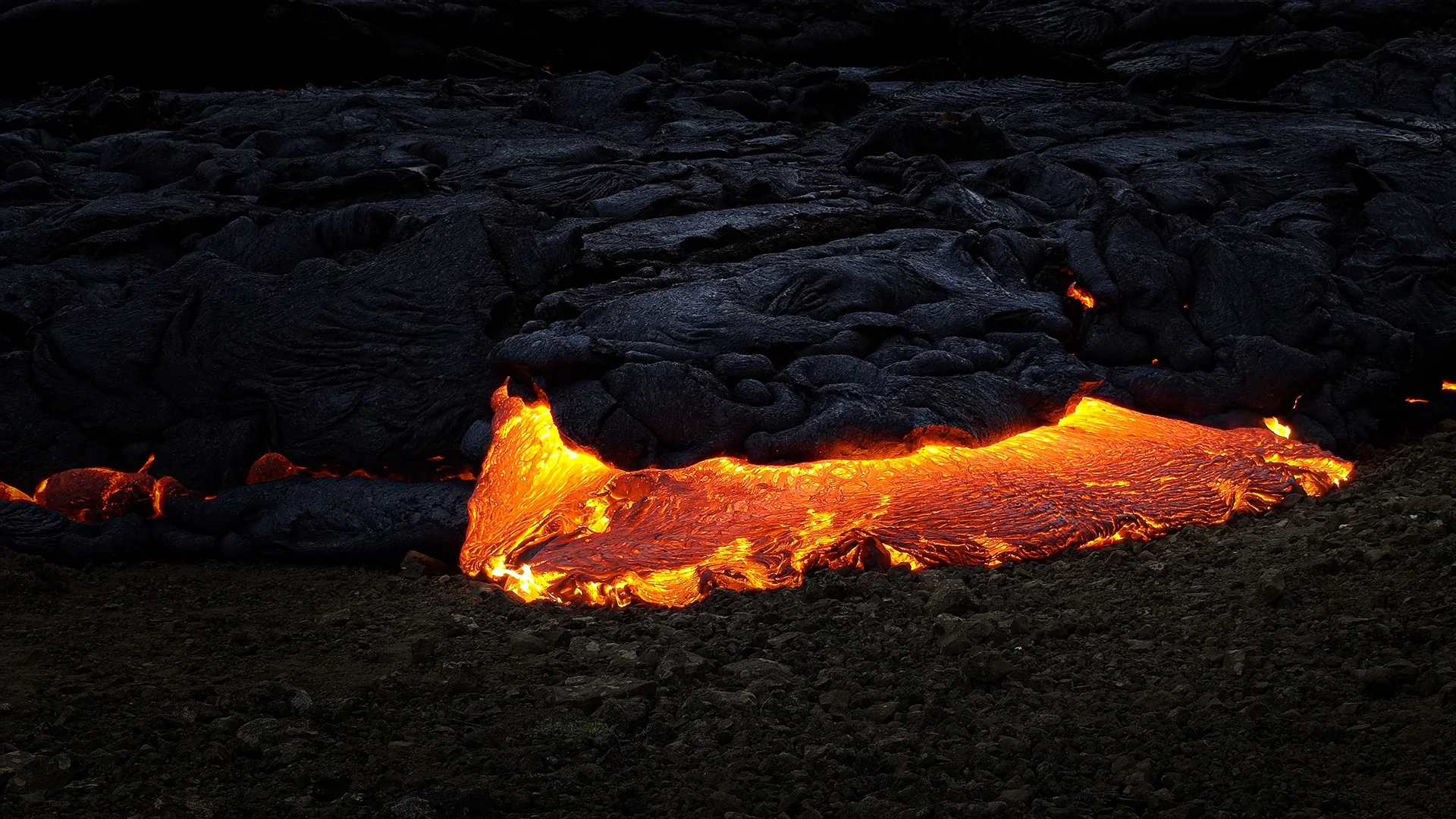 Glühend orangefarbene Lava, die unter einer Kruste aus schwarzem Gestein auf einem isländischen Lavafeld hervorfließt